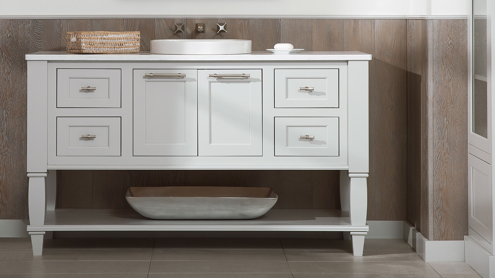 A free-standing furniture style vanity with turned posts legs on each corner and a slotted floor shelf below shown in a light gray painted finish.